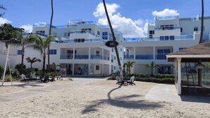 A beachfront hotel with multiple floors, palm trees, and a sandy beach area. - Beach Condo 2 Pools Walk to Beach Bars Eats (Punta Cana)