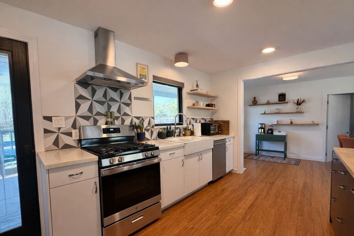 A modern kitchen with a stove, oven, and microwave. There are wooden shelves above the counter and a dining area with a table and chairs.