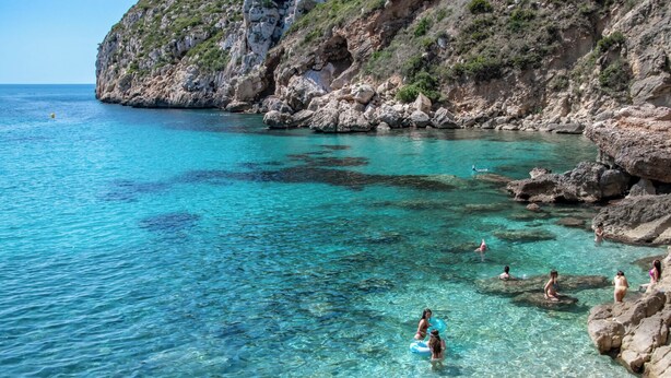 Une zone côtière caractérisée par une eau turquoise et limpide, des falaises rocheuses, et par la présence de personnes qui se baignent et profitent de la plage.