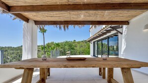 Une table en bois, sur laquelle reposent deux verres à vin, est placée sur un balcon offrant une vue dégagée sur un paysage verdoyant.
