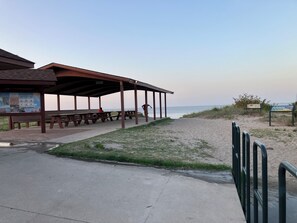 Un pabellón frente a la playa con mesas de picnic, un sendero de hormigón y una zona arenosa con un cartel.