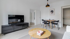 Ein modernes Wohnzimmer mit Flachbildfernseher, einem Esstisch mit Stühlen, einem Couchtisch mit Büchern und einer Pendelleuchte.
