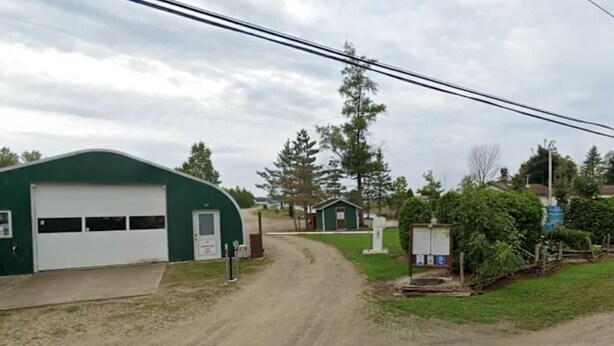 A gravel road leading to a green building with a white garage door, surrounded by trees and a small structure with a sign. - Fantastic campground retreat #35 (Lion's Head)
