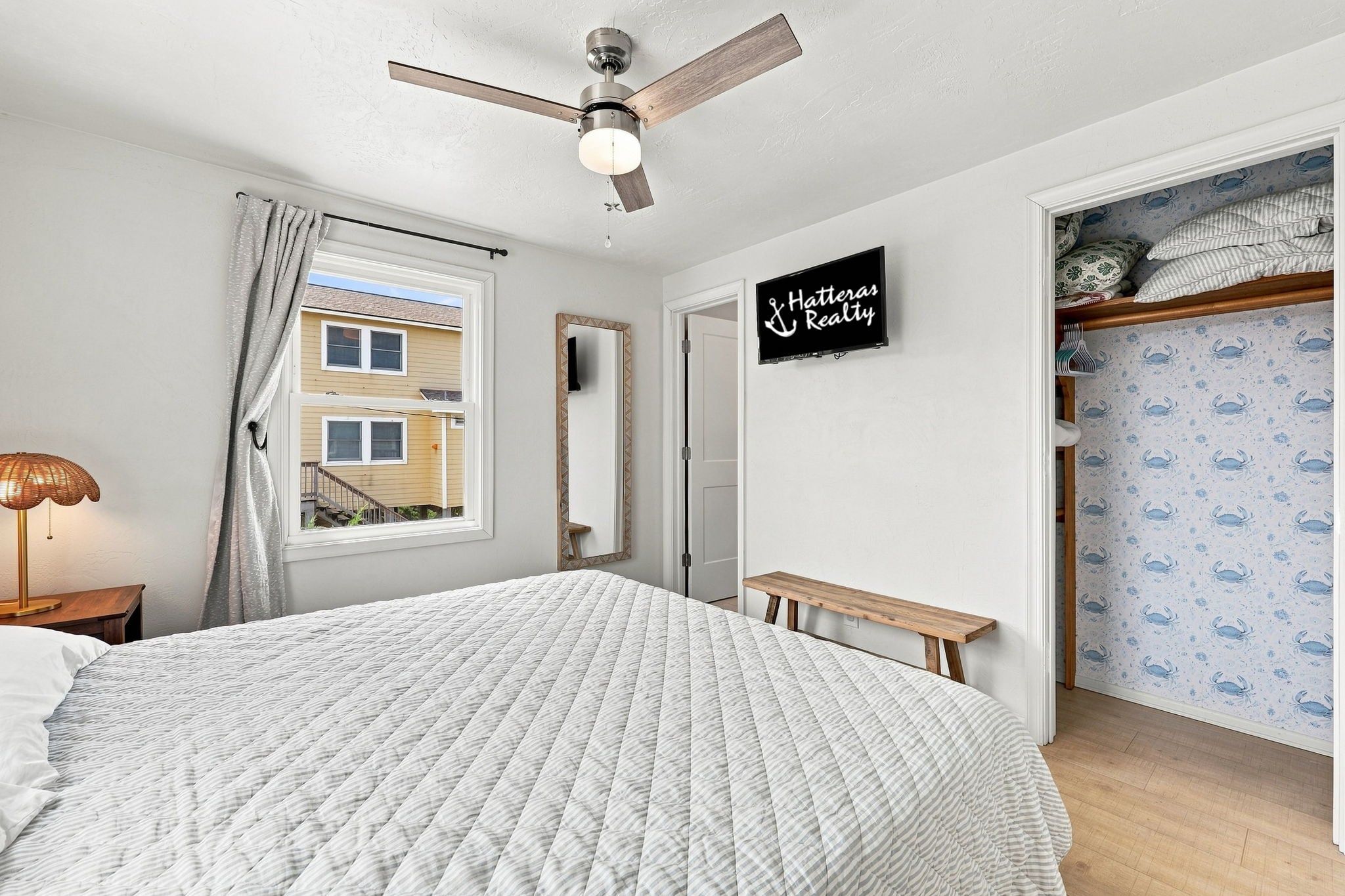 A bedroom with a bed, a ceiling fan, a window with curtains, a mirror, a bench, and a wall with a sign.
