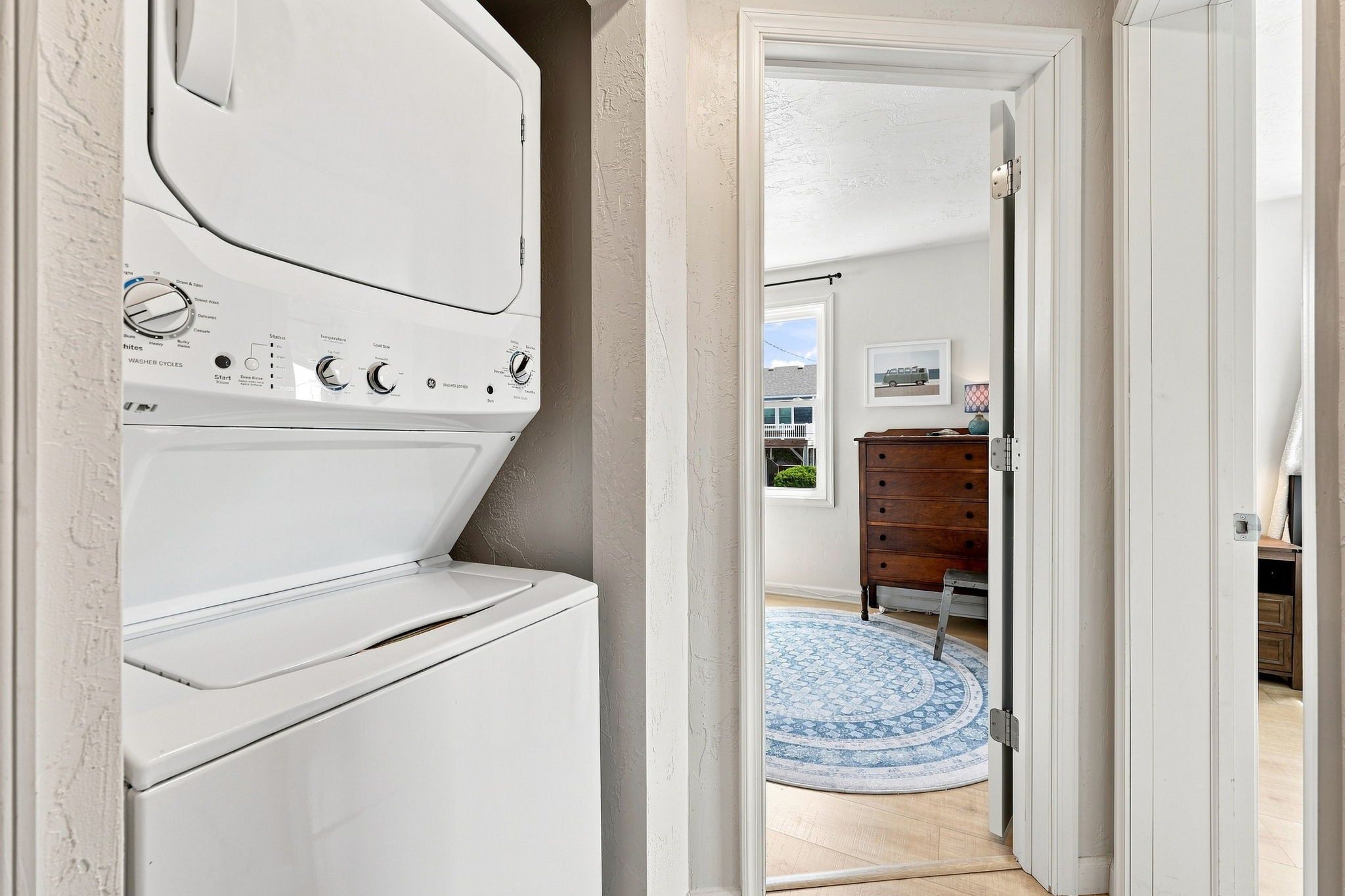 A laundry room with a front-loading washer and dryer, a wooden dresser, and a blue patterned rug in the adjacent hallway.