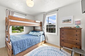 Ein Etagenbett mit blauer Steppdecke, eine Holzkommode, ein gemustertes Teppich und ein Fenster mit Blick auf ein Haus und Grünflächen.