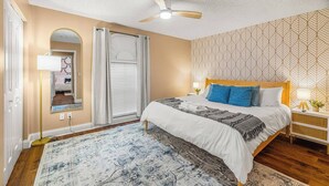 Um quarto com cama, ventoinha no teto, mesa de cabeceira, espelho, janela com cortinas e parede estampada.