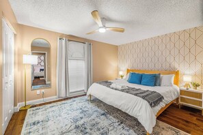 Ein Schlafzimmer mit Bett, Deckenventilator, Nachttisch, Spiegel, Fenster mit Vorhängen und einer Wand mit Muster.