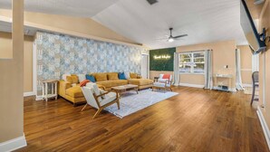 Uma sala de estar ampla, com piso de madeira, um sofá amarelo, uma poltrona branca e um ventilador de teto.
