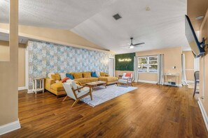 Ein geräumiges Wohnzimmer mit Holzboden, einem gelben Sofa, einem weißen Sessel und einem Deckenventilator.
