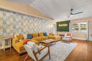Ein Wohnzimmer mit einer Wand mit Muster, einem Ecksofa, einem weißen Sessel, einem Couchtisch und einem Deckenventilator.