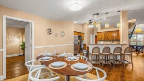 Uma sala de jantar com uma mesa redonda posta para seis pessoas, rodeada por cadeiras brancas, num espaço com piso de madeira e paredes bege.