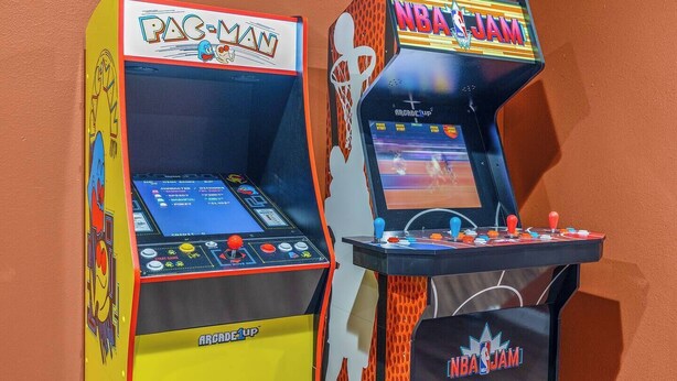 Duas máquinas de jogos de fliperama, uma com a etiqueta 'PAC-MAN' e outra com 'NBA JAM', ambas com gráficos coloridos e controles de joystick.