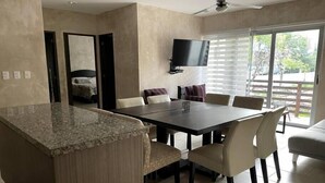 A modern kitchen with granite countertops, a dining area with a dark table and beige chairs, a flat-screen TV mounted on the wall, and a large window with blinds. - Carmela  (Tulum)