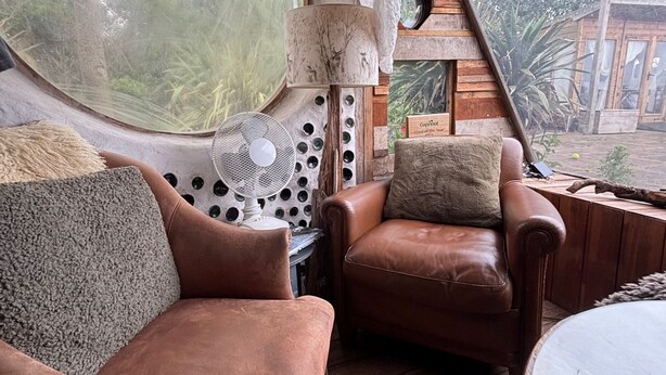 Interior acolhedor de uma cabana, com uma janela redonda, um ventilador, um sofá de couro, uma mesa de madeira e vista para o exterior.