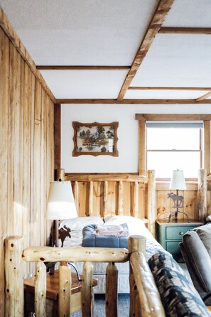 A rustic wooden room with a bed, a wooden bench, a lamp, and a framed picture on the wall.