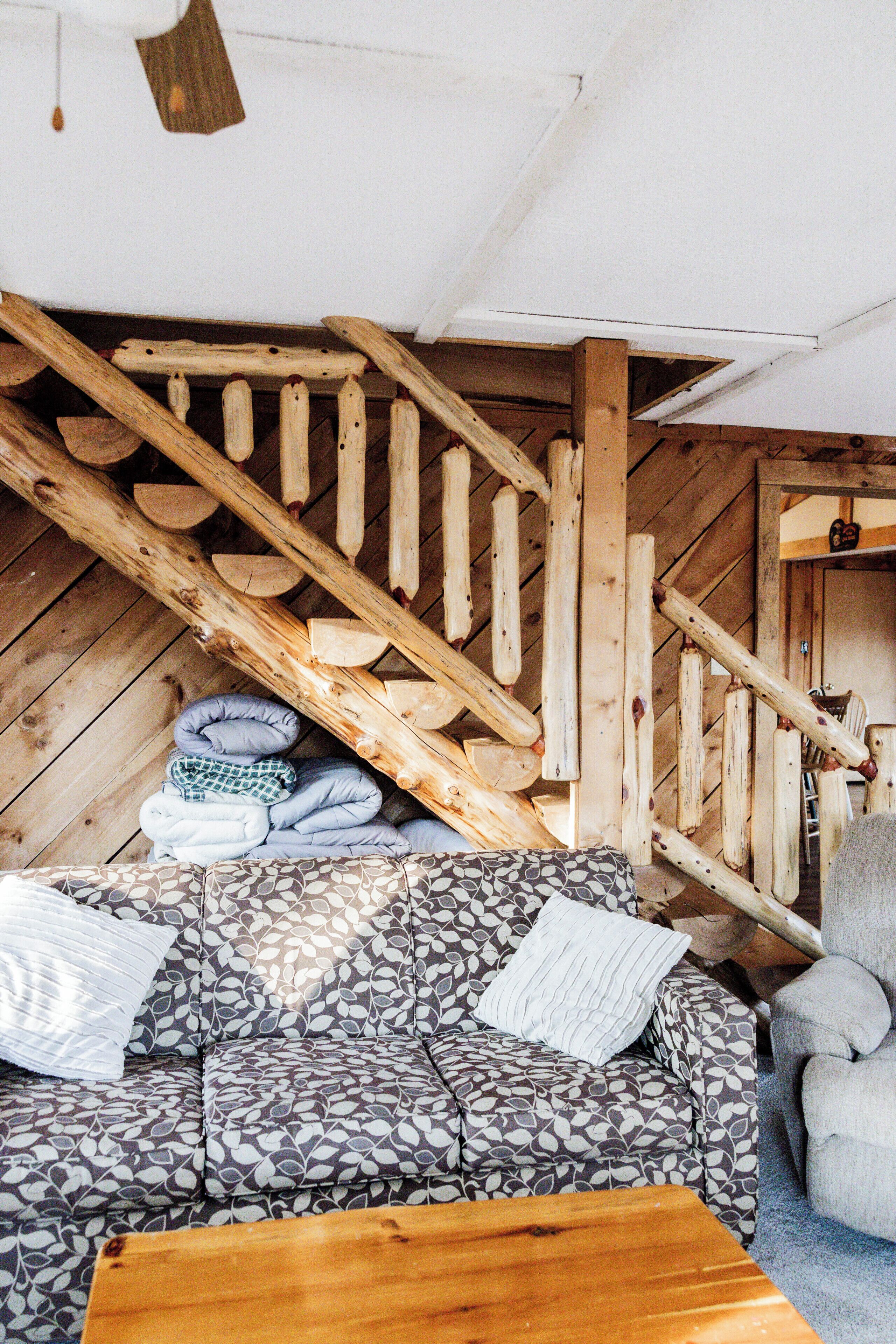 A cozy cabin interior with a wooden staircase, a patterned sofa, and a chair.