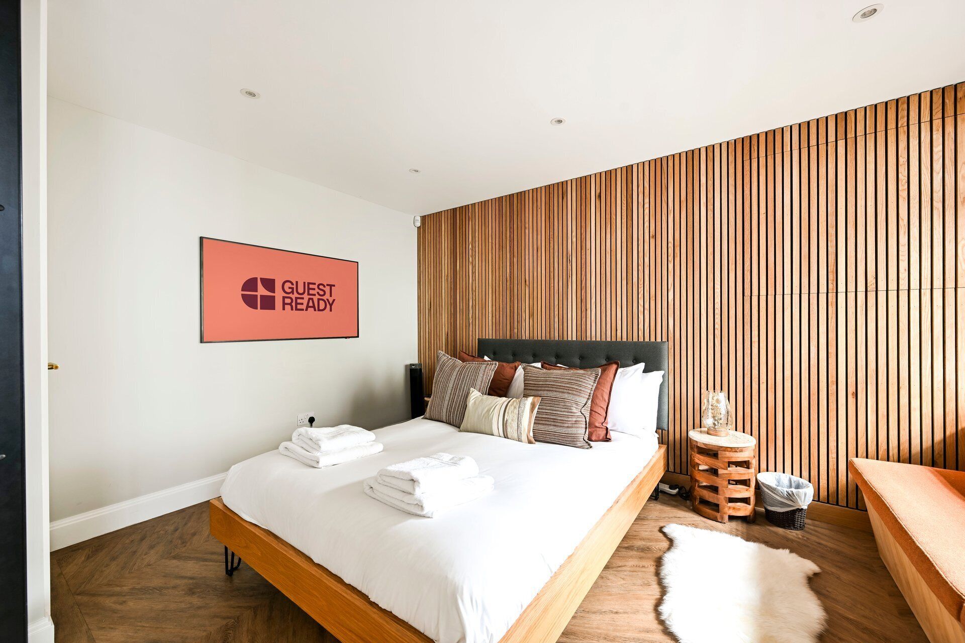 A modern bedroom with a bed, pillows, a wooden headboard, a 'GUEST READY' sign, a small wooden stool, and a white fur rug.