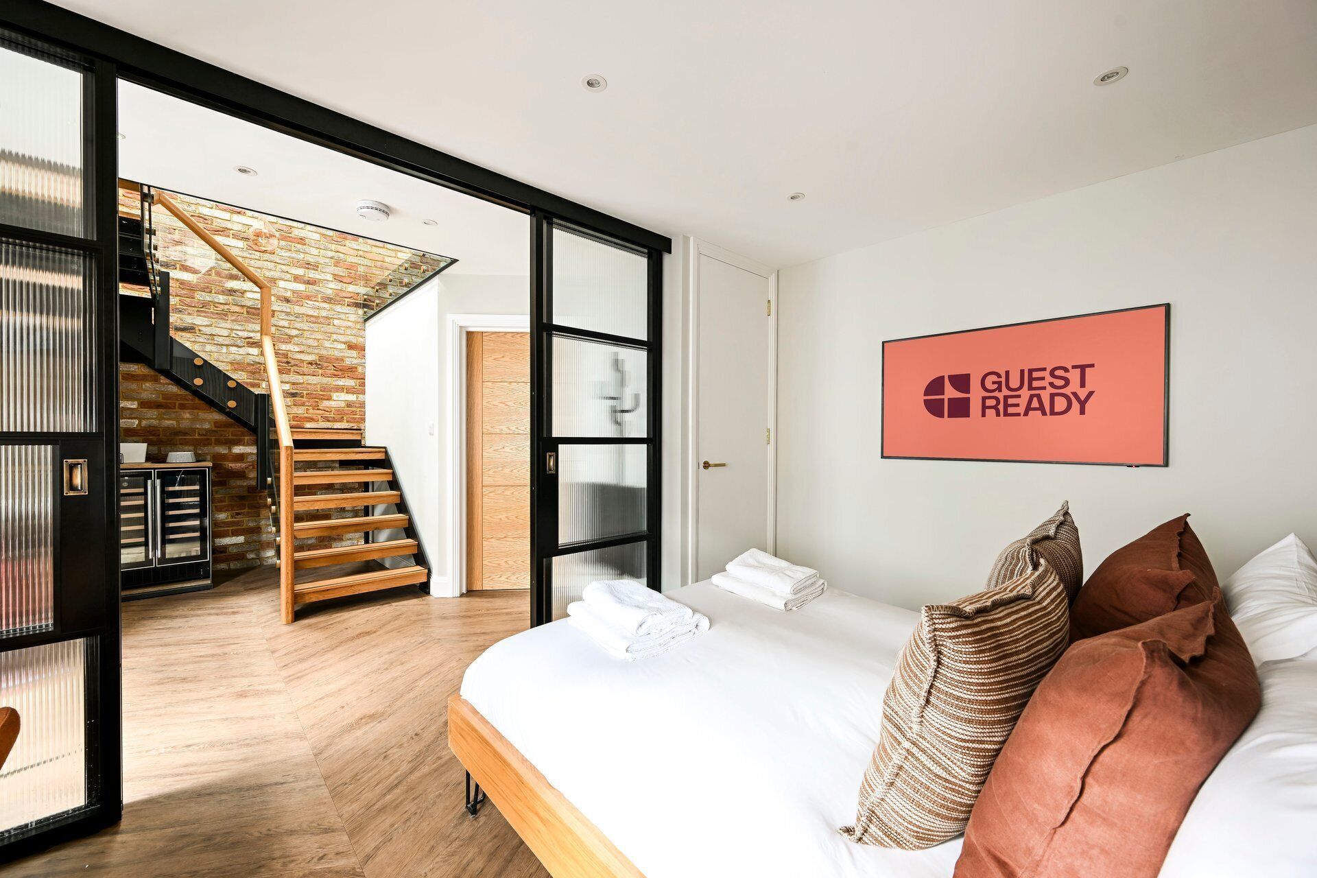 A hotel room with a bed, pillows, a 'GUEST READY' sign, a staircase, and a view of a brick wall.