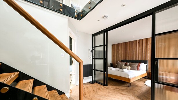 Een moderne hotelkamer met een houten vloer, een bed met veel kussens, een glazen deur naar een balkon en een trap met houten treden.