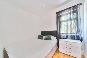 A bedroom with a bed, a bedside table, a lamp, and a window with sheer curtains.