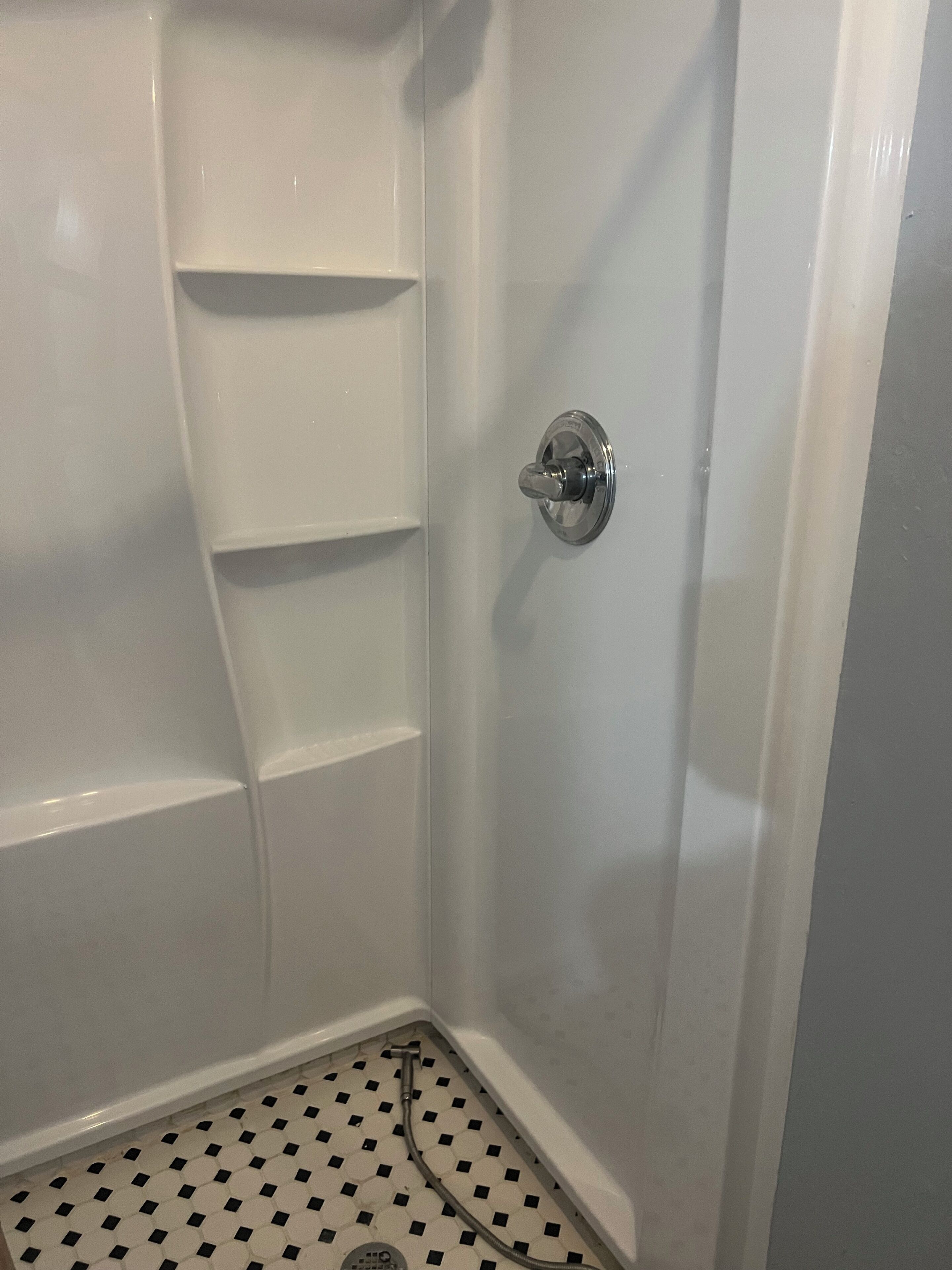 A shower with a white tiled floor, a chrome shower knob, and a wall-mounted shelf.