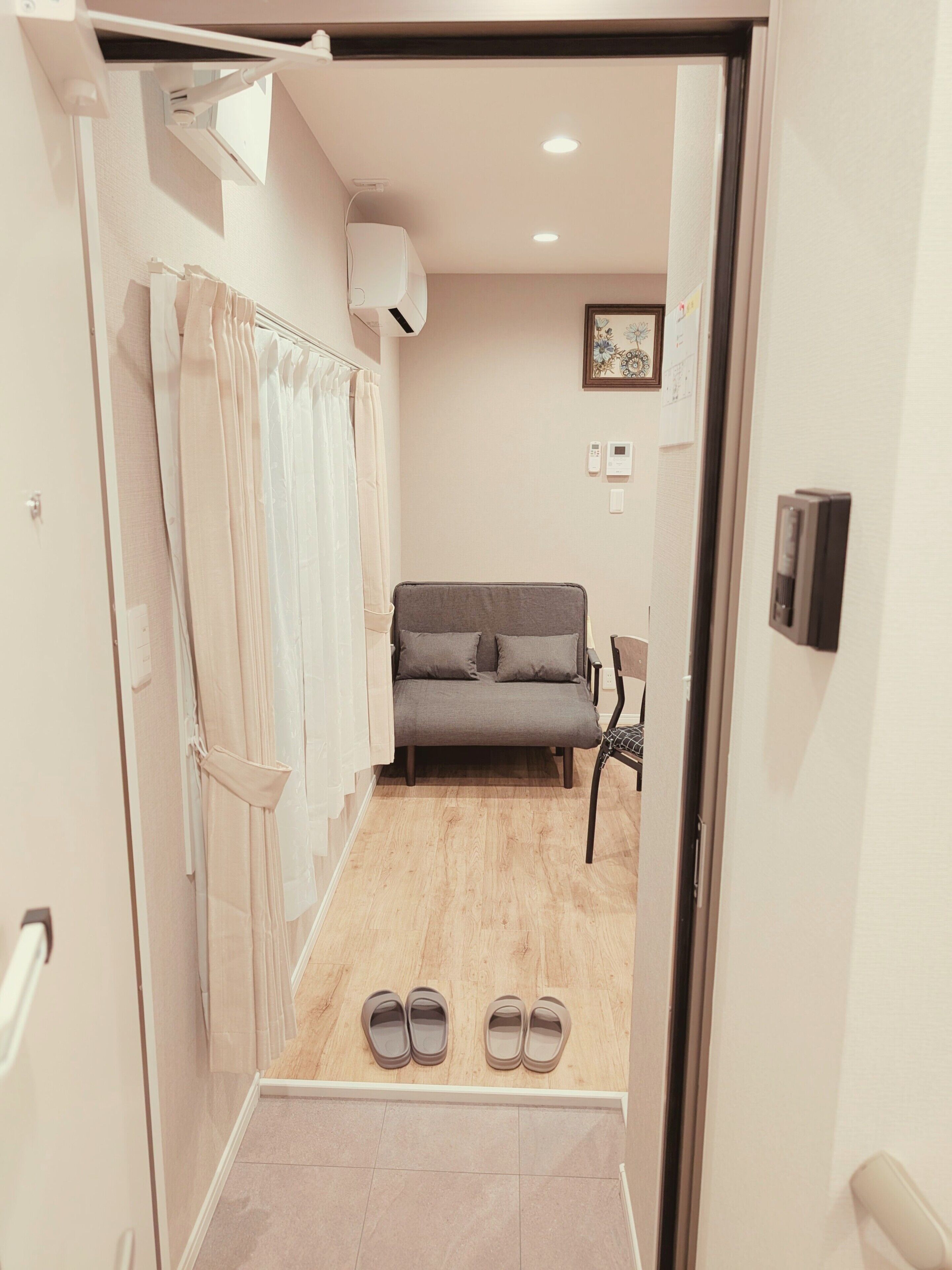 Una habitación con sofá, una silla, un par de pantuflas en el suelo, una cortina y aire acondicionado de pared.