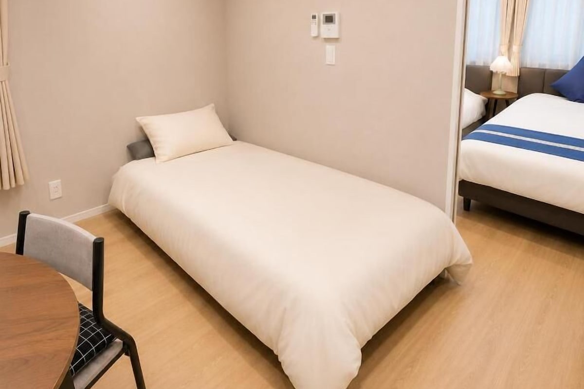 A single bed with a white duvet, a small table with a chair, an air conditioning unit, a wall-mounted phone, and a painting.