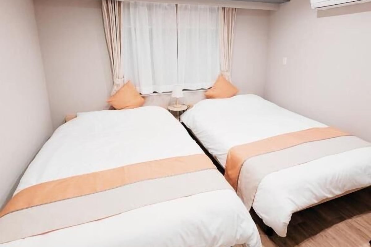 A room with two beds, a window with curtains, an air conditioning unit, and a ceiling light.
