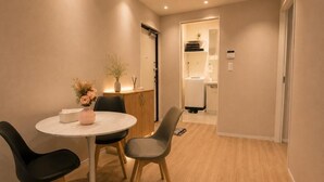 Et moderne hotelværelse med et rundt spisebord, to stole, en lille vase med blomster, et skab og et badeværelse, der ses gennem en åben dør.