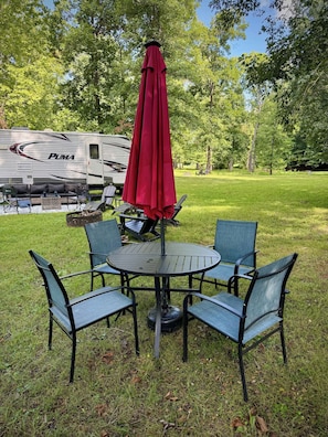 Een picknicktafel met vier stoelen en een rode parasol in een gazon met bomen.