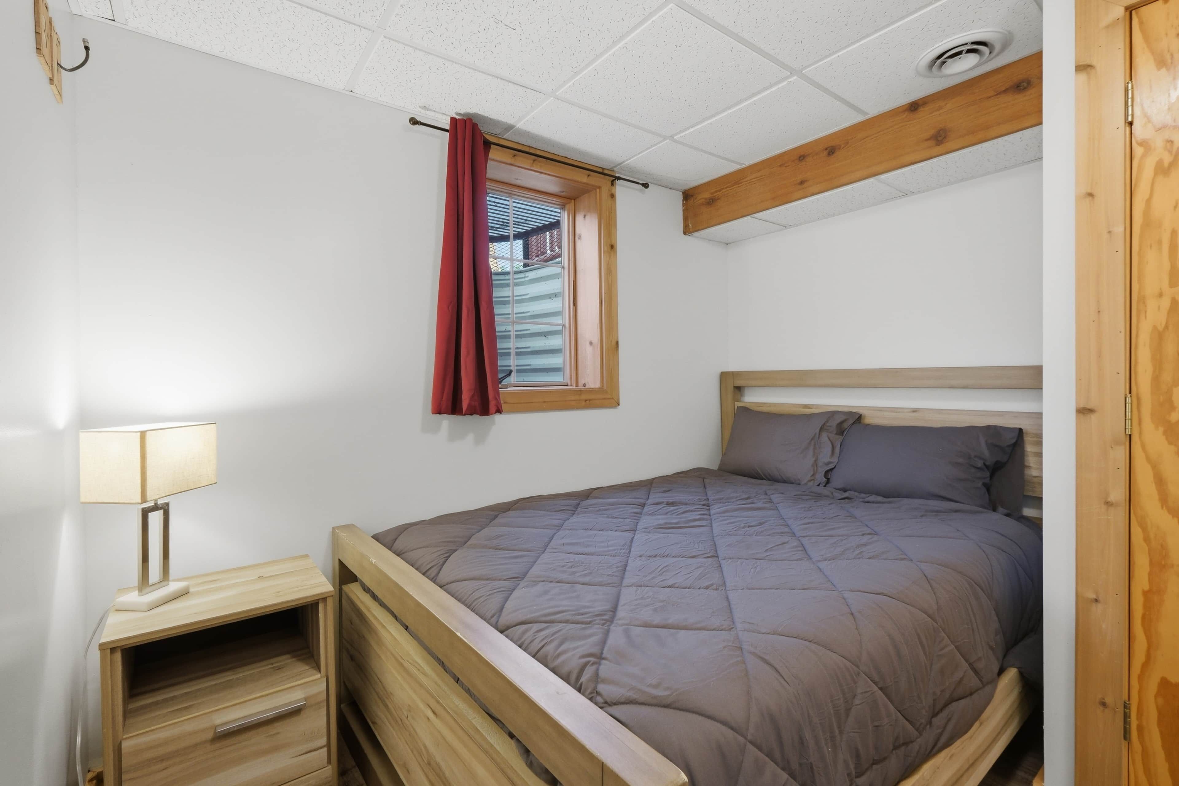 A single bed with a wooden headboard, a bedside table with a lamp, a window with a curtain, and a ceiling with exposed beams.