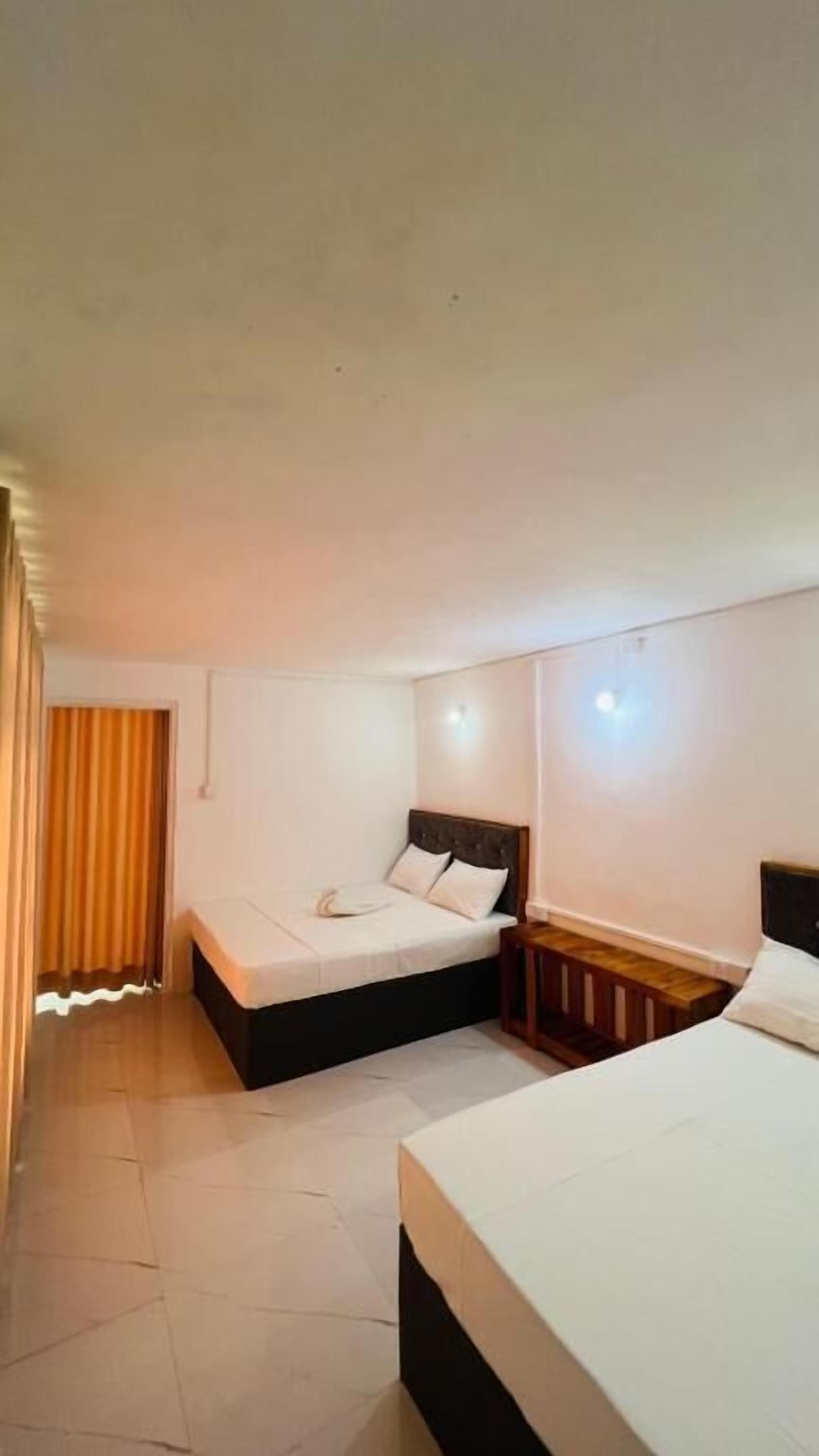 Quarto de hotel com duas camas, mesa de cabeceira, luminária na parede e cortina.