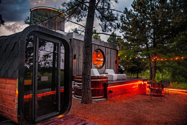 A modern outdoor cabin with a circular window, wooden deck, and a seating area with a fire pit.