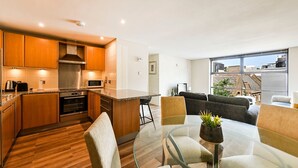 Una cocina moderna con armarios de madera, una campana extractora de acero inoxidable y un microondas. Hay una mesa de comedor de vidrio con sillas y una pequeña planta en maceta. La habitación tiene un ventanal con vistas a edificios.