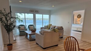 Uma sala de estar com vista para a água, uma placa com a inscrição 'Beach House' e uma área de jantar com mesa e cadeiras de madeira.