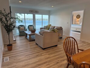 A living room with a view of the water, a sign reading 'Beach House,' and a dining area with a wooden table and chairs.