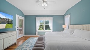 Um quarto com cama, televisão, ventoinha no teto, janela com cortinas e porta.