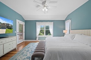 A bedroom with a bed, a TV, a ceiling fan, a window with curtains, and a door.
