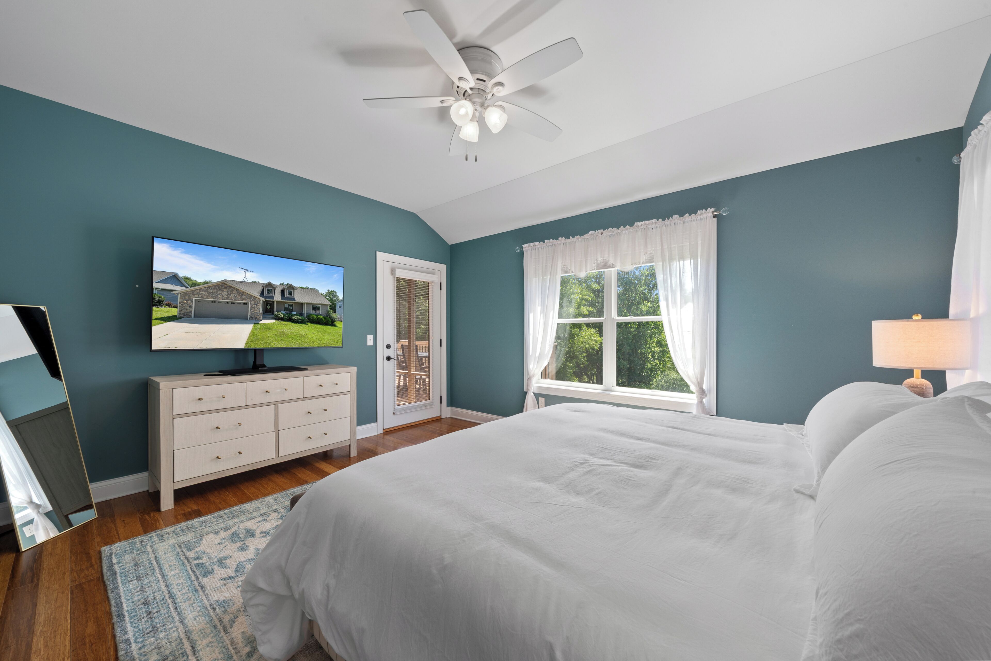 Une chambre à coucher avec un lit, un ventilateur de plafond, un téléviseur posé sur une commode, une fenêtre avec des rideaux et une vue sur une maison à l’extérieur.
