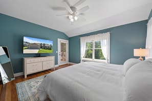 Un dormitorio con cama, ventilador de techo, televisor sobre una cómoda, ventana con cortinas y vista a una casa exterior.