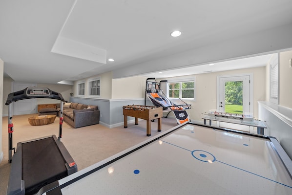 Una habitación con mesa de fútbol de mesa, cinta de correr, sofá y ventana con vista a follaje.