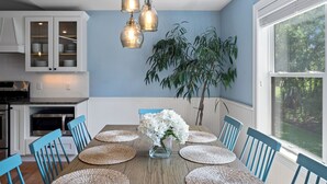 Uma sala de jantar com mesa de madeira, cadeiras azuis, um vaso com flores e luminárias pendentes.