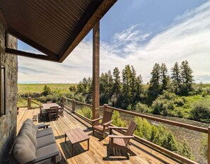 A wooden deck with outdoor furniture, a stone wall, and a view of a river and forest. - Luxury Riverfront 5BR Lodge with hot tub, fireplace, custom bar, and mountain views. (Ashton)