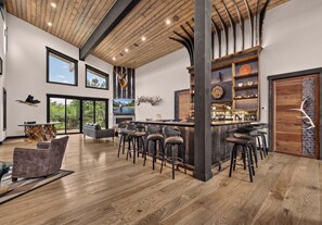 A spacious interior with a wooden ceiling, large windows, a bar area with stools, and a fireplace. - Riverfront 5BR lodge with hot tub, fireplace, and mountain views. (Ashton)