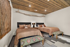 A bedroom with two beds, a wooden ceiling, a wall-mounted artwork, and a patterned rug. - Riverfront 5BR lodge with hot tub, fireplace, and mountain views. (Ashton)
