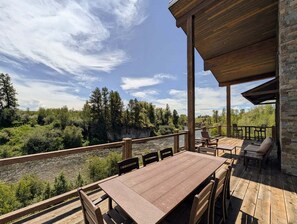 A wooden deck with a table and chairs overlooking a river and forest. - Riverfront 5BR lodge with hot tub, fireplace, and mountain views. (Ashton)