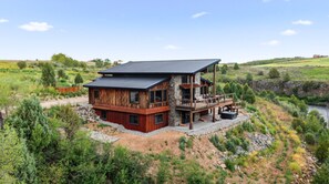 A two-story wooden cabin with a balcony, surrounded by natural landscape. - Luxury Riverfront 5BR Lodge with hot tub, fireplace, custom bar, and mountain views. (Ashton)