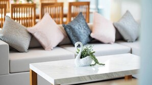 Ein modernes Wohnzimmer mit einem weißen Sofa, grau und rosa Kissen, einer weißen, schwanförmigen Vase mit Grünpflanzen und einem Holztisch.