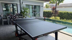 Ein Tischtennis-Tisch auf Rädern, ein Ball und ein Schläger, platziert auf einer Terrasse mit Schwimmbecken im Hintergrund.